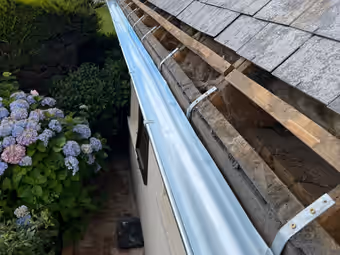 Gouttière en zinc demi-ronde neuve fixée en rive d’une toiture ancienne, un massif d’hortensias en contrebas